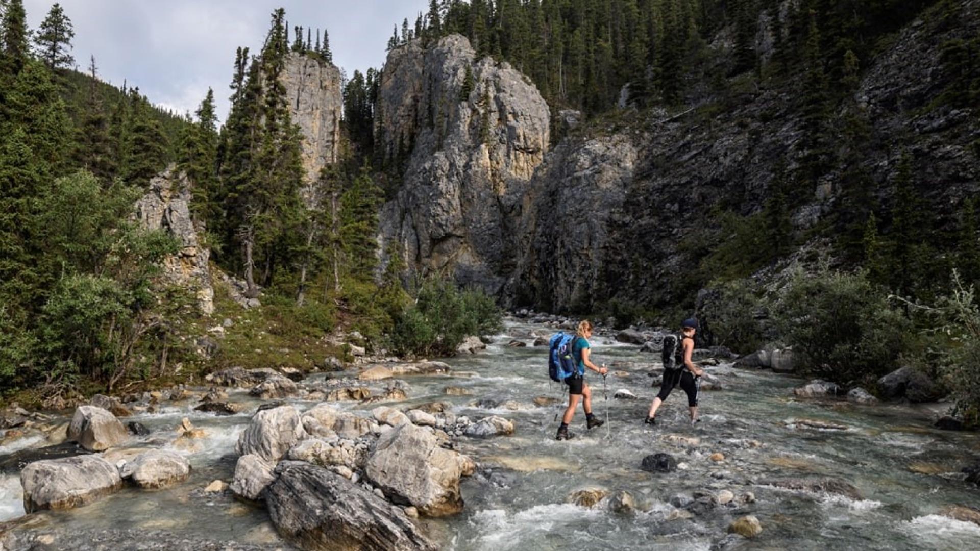Two hikers crossing a rocky stream surrounded by forested mountains.