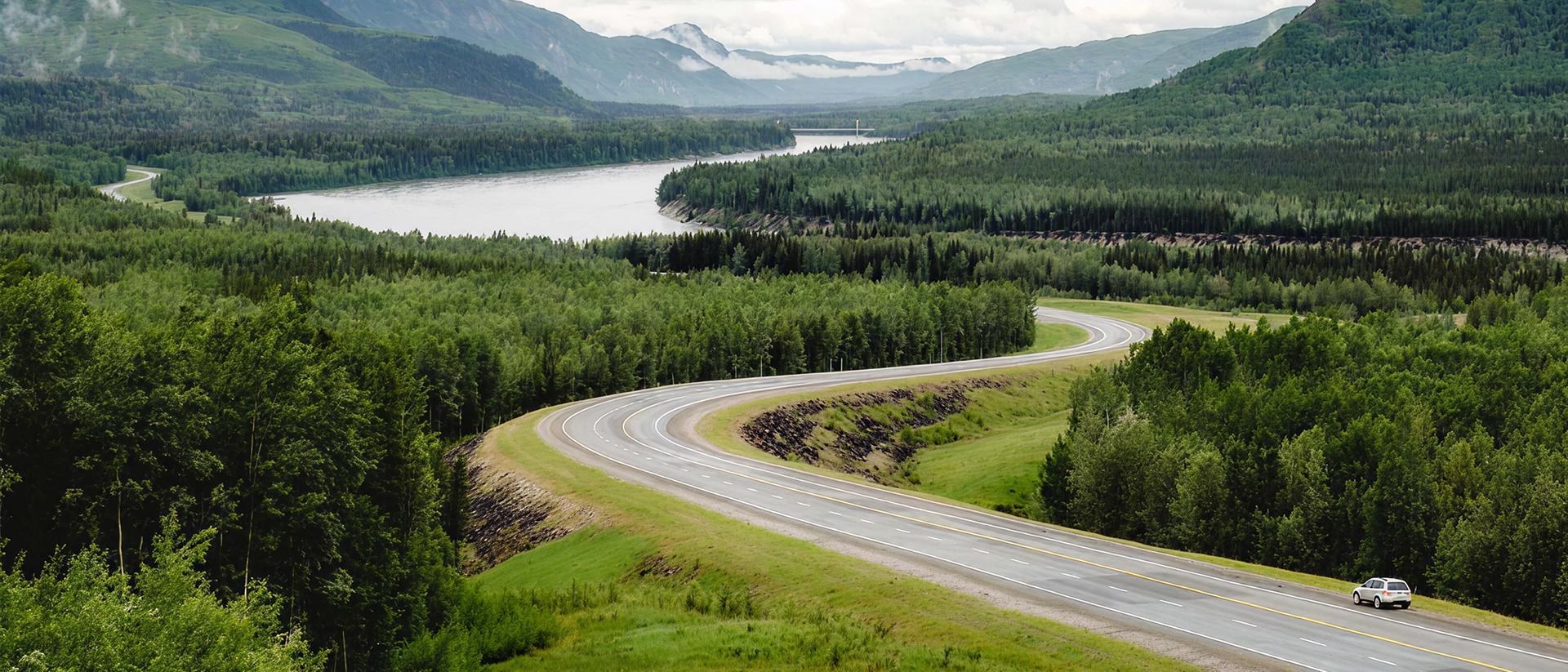 Winding road through lush green forest near a river and mountains.