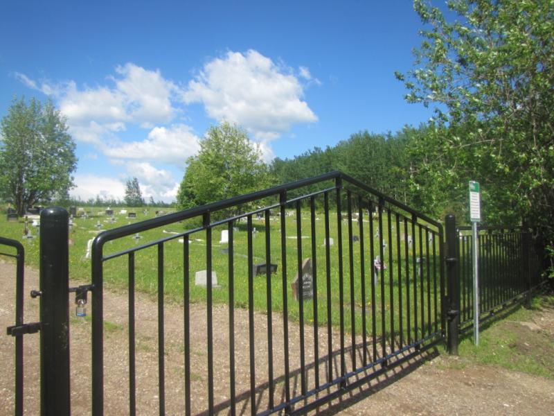 Black metal gate at a cemetery entrance on a sunny day with trees and tombstones.