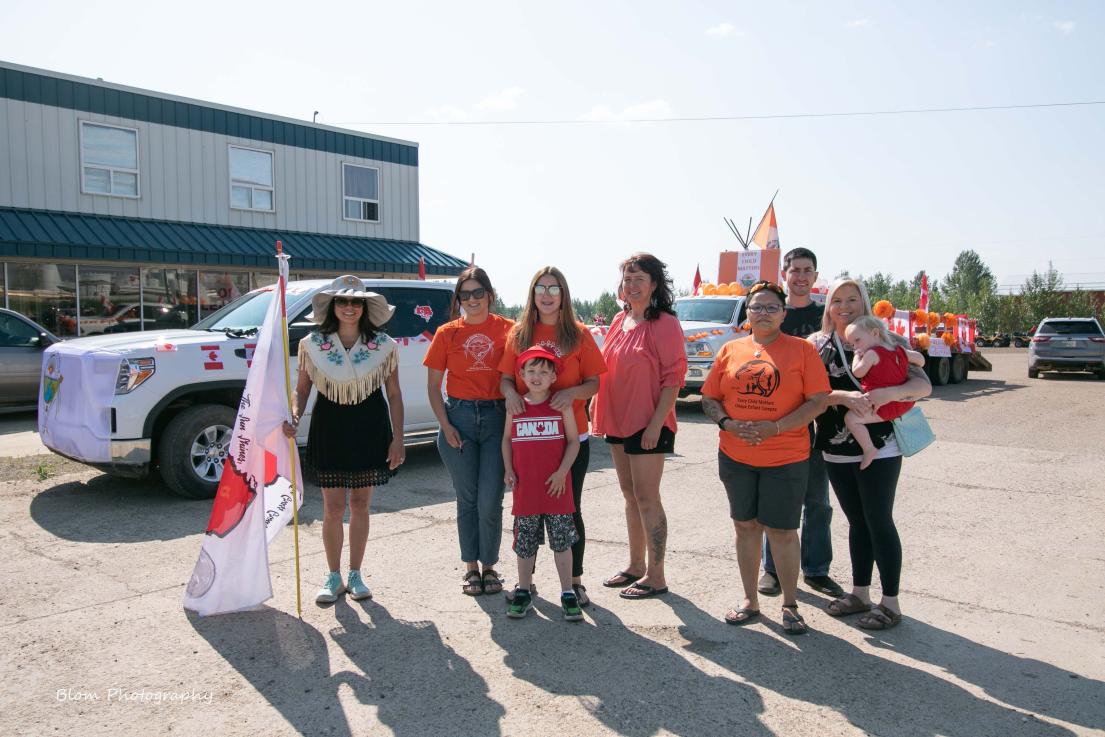 People in orange shirts and one in traditional attire at an outdoor gathering near vehicles.