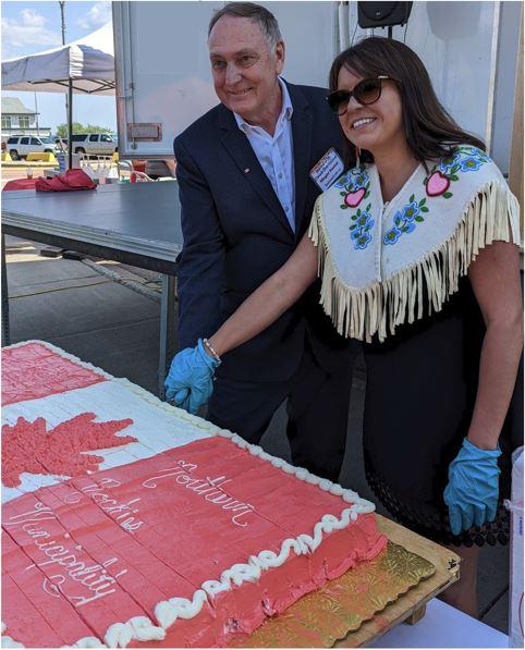 Two people cut a large red and white cake outdoors.