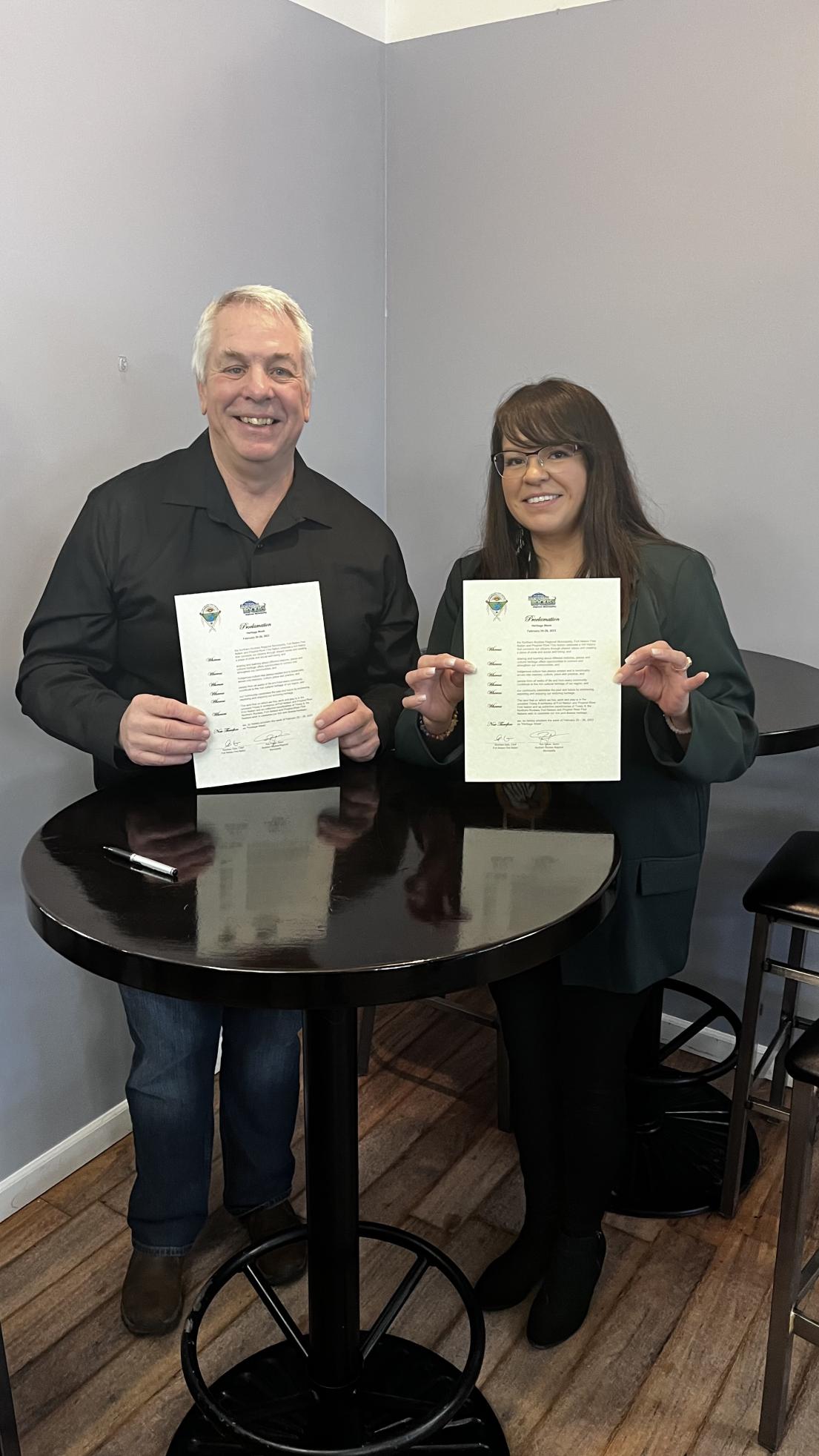 Two people smiling, holding certificates at a round table.