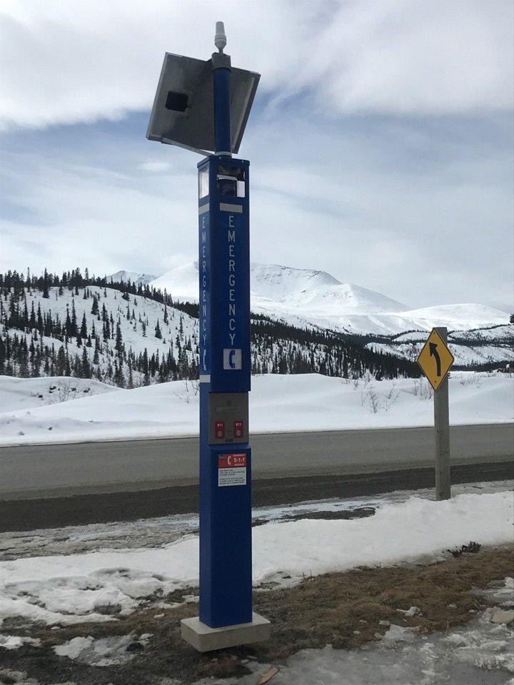 Blue weather station signpost by a snowy roadside, mountains in the background.